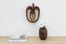 Decorative mask on a wall above a vase with flowers and books on a wooden surface.
