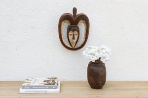 Decorative mask on a wall above a vase with flowers and books on a wooden surface.