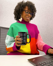A woman with curly hair and glasses wearing a vibrant multicolor color-block sweater, holding a black mug with white geometric patterns while sitting at a desk with a laptop.