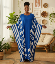 Woman wearing a blue and white patterned kaftan dress in a room with plants and decor.