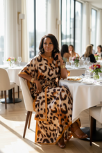 Woman in a leopard print kaftan dress sitting at a table in a bright room with large windows.