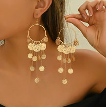 Gold hoop earrings with dangling coins being worn by a person.
