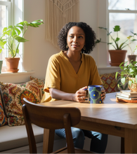 A woman with curly black hair sits at a wooden table in a bright, sunlit room, holding a blue and yellow patterned mug. She is wearing a mustard yellow V-neck top.