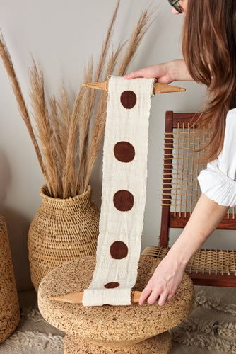 Person holding a decorative fabric strip with brown circles on a woven stool.