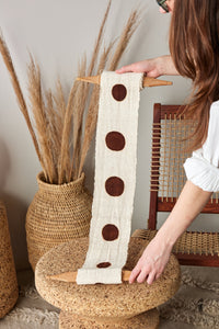 Person holding a decorative fabric strip with brown circles on a woven stool.