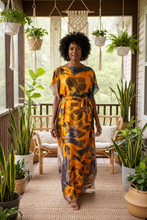 Woman in a orange floral kaftan dress standing on a porch with plants