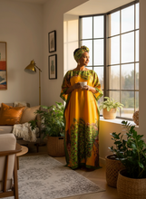 Woman in a orange kaftan dress standing in a sunlit living room with plants and decor.