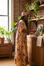 Woman in a yellow dress standing in a kitchen with plants and wooden cabinets.