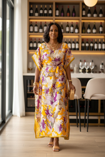 Woman in a yellow floral kaftan dress standing in a modern wine bar with wine bottles on shelves.