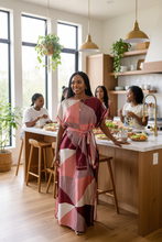 Woman in a pink patterned kaftan dress standing in a modern kitchen with friends around a dining table.