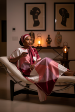 Woman in a pink kaftan dress sitting on a chaise in a room with framed art and decorative items.