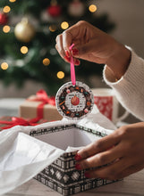 Person holding a decorative pink ornament with a Christmas tree and wrapped gifts in the background