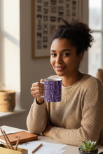 A woman with curly hair sitting at a desk, holding a purple and white patterned ceramic mug. She is wearing a beige knit sweater and smiling.