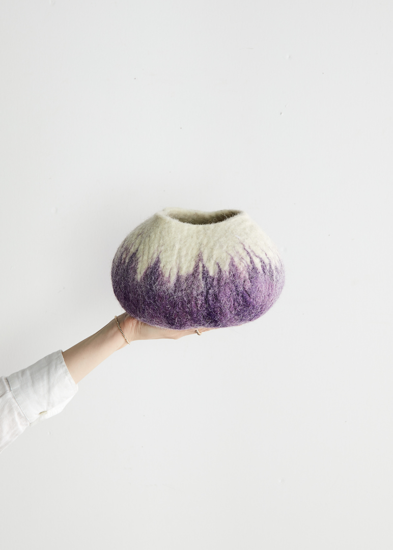 Hand holding a textured wool vase with a gradient from purple to beige on a light gray background
