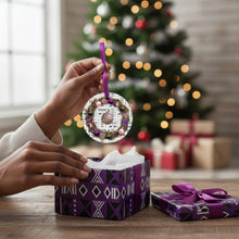 Person holding a Christmas ornament with a decorated tree and presents in the background