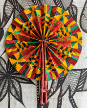 A round folding hand fan with a vibrant Kente-style geometric pattern in yellow, red, green, and black, featuring natural leather handles tied together.