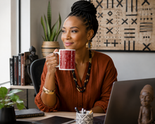 A woman with braided hair sits at a desk holding a red and white patterned ceramic mug. She is wearing a rust-colored blouse and jewelry, with a laptop, books, and plants in a modern office setting.