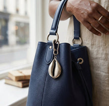 Navy blue leather handbag with a cowrie shell charm held by a person indoors.