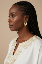 Woman with braided hair wearing a white top with gold embroidery on a beige background