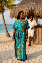 Woman in a blue kaftan dress standing on a beach with a thatched-roof hut and palm tree in the background.