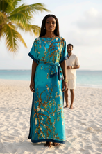 Woman in a blue floral kaftan dress standing on a beach with palm trees with her husband in the background.