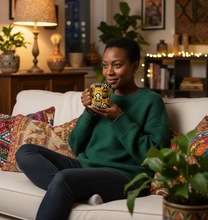 A woman sits on a white sofa in a cozy, warmly lit living room, holding a square ceramic mug with a colorful African wax print pattern featuring black, yellow, and white geometric designs.