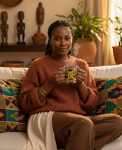 A woman sits on a white sofa in a warm, decorated living room, holding a yellow and black patterned mug. She is wearing a rust-orange knit sweater and brown pants, with colorful patterned cushions behind her and decorative wall art in the background.