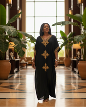 Woman in a black dress with gold patterns standing in a luxurious interior setting.
