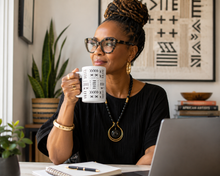 A woman with braided hair and glasses sits at a desk holding a white mug with black geometric patterns. She wears a black pleated top and a black beaded necklace with a gold-toned pendant.