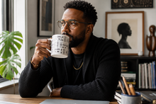 A man with glasses and a beard sits at a desk drinking from a white ceramic mug with a black geometric pattern. He is wearing a black ribbed sweater in a home office setting with framed art, books, and a plant in the background.