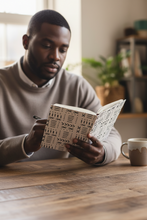 A man in a beige sweater sits at a wooden table, reading a notebook with a black and white geometric pattern. A ceramic mug is on the table next to him, with a softly blurred indoor background.
