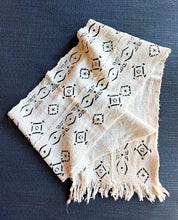 A square cream-colored woven scarf with black geometric and abstract patterns, featuring fringed edges, laid flat on a dark textured blue background.