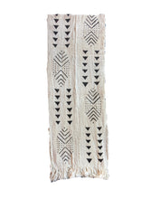 A rectangular woven textile with fringe at the bottom, featuring black geometric tribal-style patterns on a warm off-white background.
