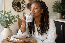 A woman with long black locs sits at a table in a bright kitchen, holding a white ceramic mug with a black geometric pattern. She is wearing a white blouse and gold jewelry.
