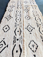 A close-up of a woven textile runner in natural beige with black geometric tribal patterns, laid flat on a dark gray surface.