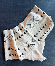 A folded cream-colored woven scarf with black geometric patterns, including triangles, chevrons, and dots, displayed on a dark textured blue background.