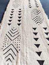 A close-up of a woven table runner with a cream base and black geometric patterns, including chevrons, triangles, and dotted lines, laid flat on a dark blue surface.