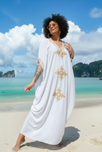 Woman in a white dress with gold patterns standing on a beach with turquoise water and blue sky.