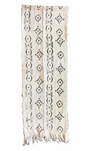 A vertical woven textile runner or scarf in a warm cream or off-white color, featuring a black geometric pattern of diamonds, crosses, and vertical lines, finished with fringed edges at the bottom.