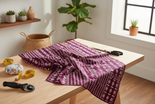 Purple patterned African fabric on a wooden table with sewing tools, plants, and a window in the background.