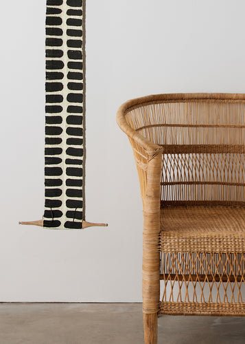 Woven chair next to a black and white patterned wall hanging on a plain wall.