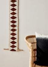 Decorative wall hanging with geometric pattern next to a wicker chair and black cushion.