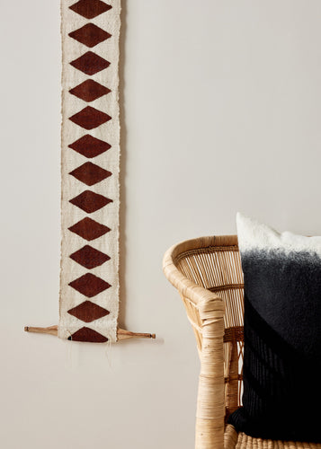 Decorative wall hanging with geometric pattern next to a wicker chair and black cushion.