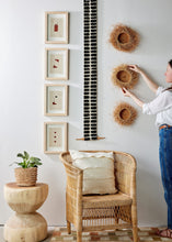 Person arranging decorative items on a wall with a wicker chair and plant in the foreground.