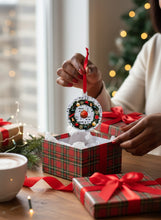 Person holding a Christmas ornament with a wreath design, surrounded by gift boxes and a tree in the background.