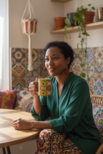 A woman in a green blouse and patterned pants sits at a wooden table holding a yellow and orange owl-themed ceramic mug in a cozy, decorated room.