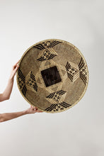 Person holding a woven round mat with geometric patterns on a plain background