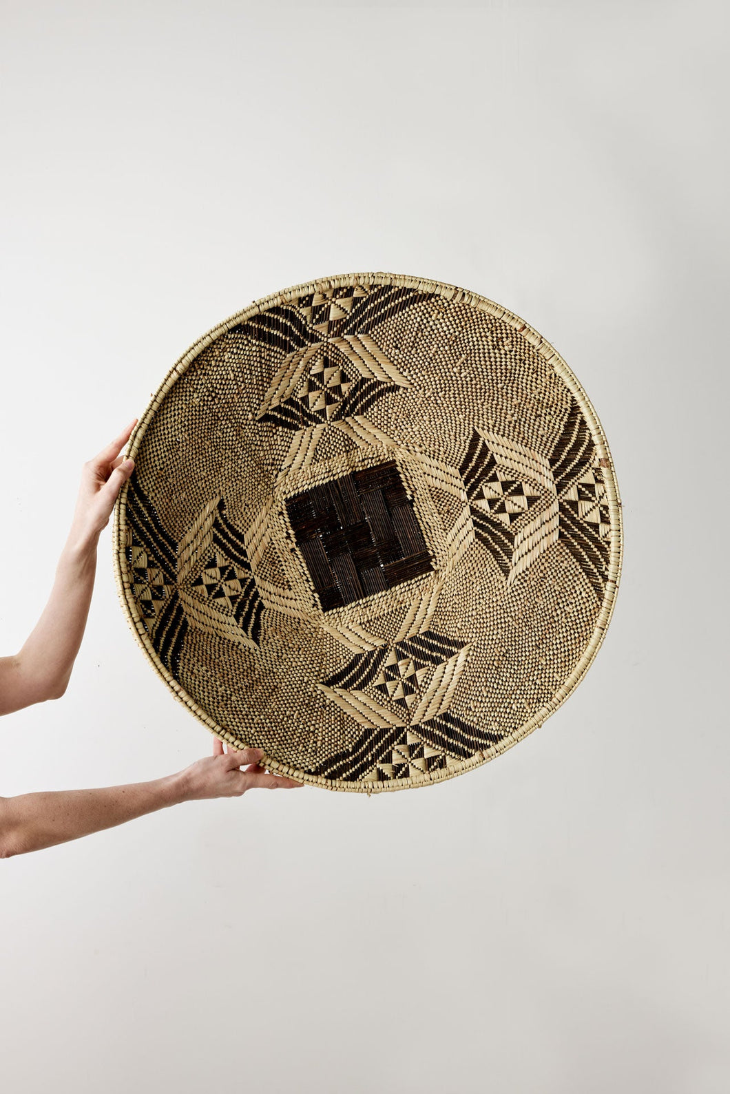 Person holding a woven round mat with geometric patterns on a plain background